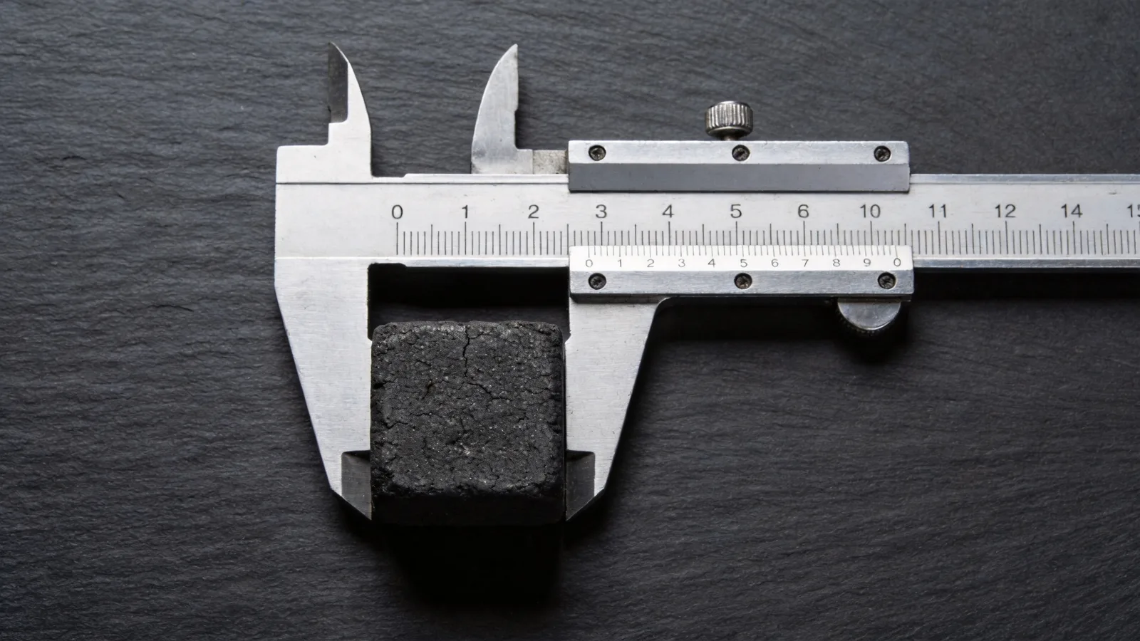 A charcoal briquette measured with a steel caliper on a slate surface — conceptual illustration